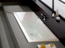 Rechteckige Badewanne aus Stahl-Email, teilweise mit Wasser gefüllt, moderner Wasserzulauf, umgeben von grauen Fliesen.